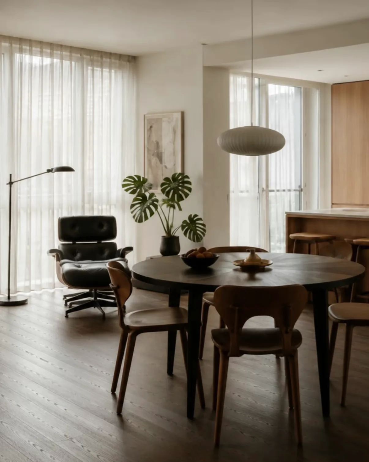 A round dark dining table with wooden chairs, generous floor space around it and sheer curtains behind.