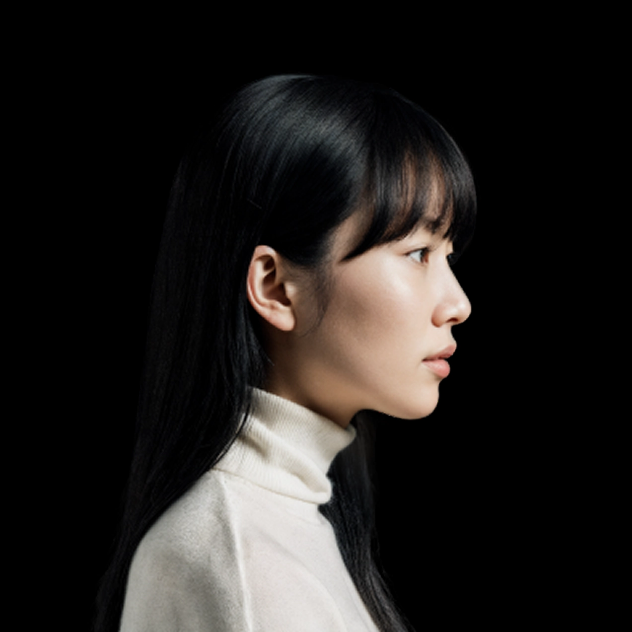 Low resolution of a profile portrait of a person with long straight black hair and bangs, wearing a cream-colored turtleneck sweater. The subject faces left against a plain black background, with soft, even lighting highlighting the side of the face and hair.